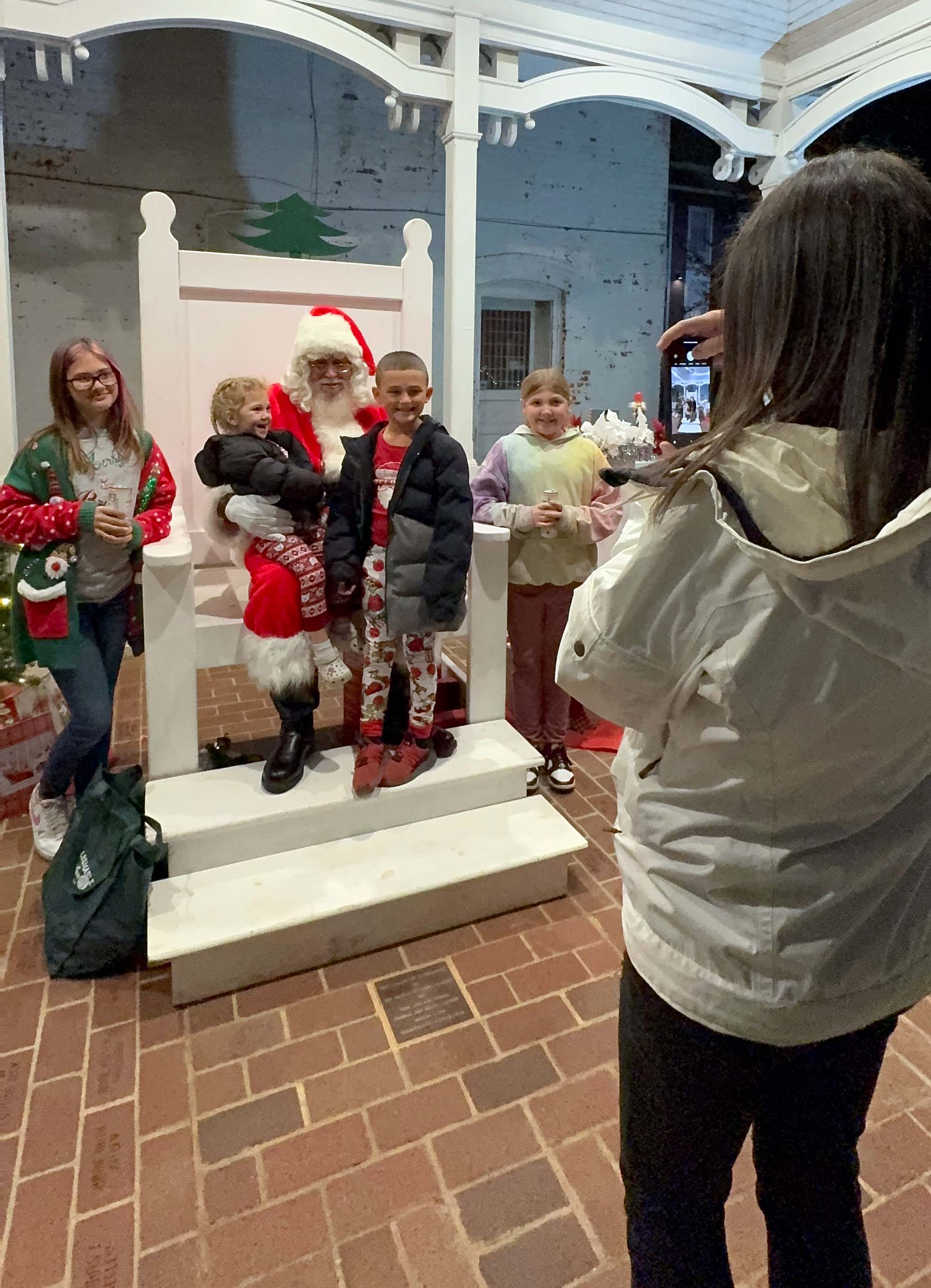 A group of children pose for a photo with Santa Claus sitting on a white chair under a gazebo. One child is sitting on Santa's lap while two others stand beside him smiling, and a woman in a white coat takes their picture with a phone. The scene is set on a brick patio with holiday decorations.